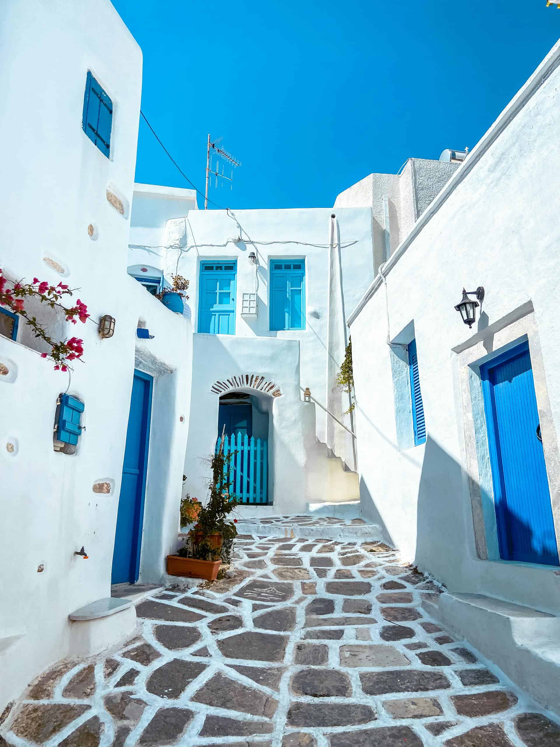 Explore a scenic Greek alley featuring blue doors and whitewashed houses under a bright blue sky.