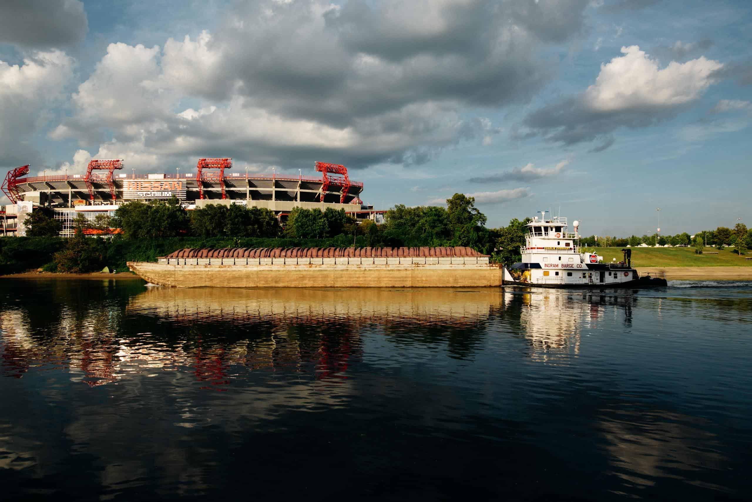 A serene view of a riverboat passing by a sports stadium with reflections, captured at sunset.
