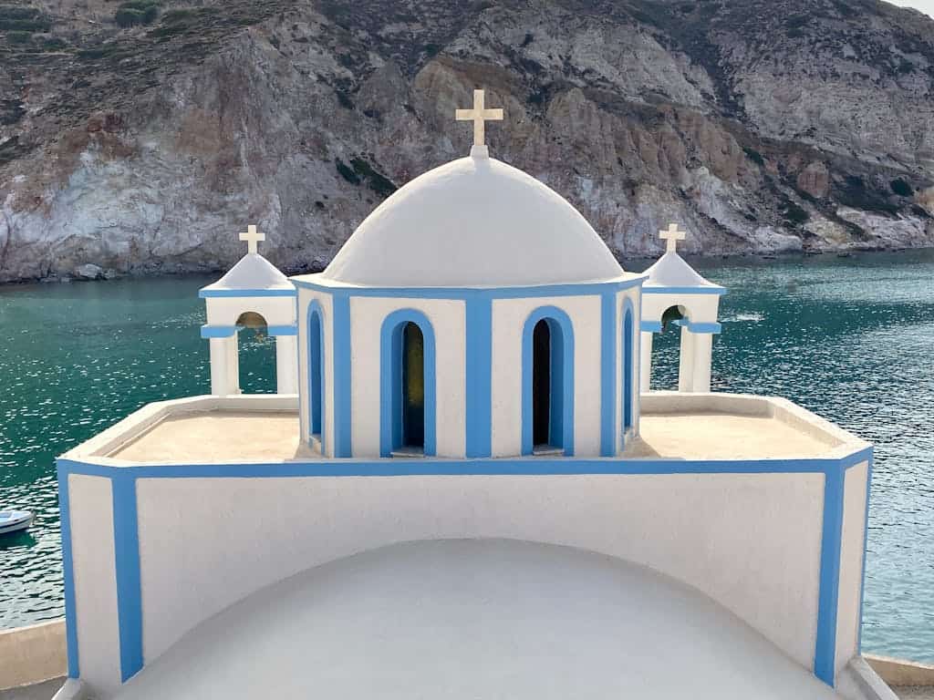 Charming white and blue chapel on Milos, Greece, by the Aegean Sea with stunning mountain backdrop.