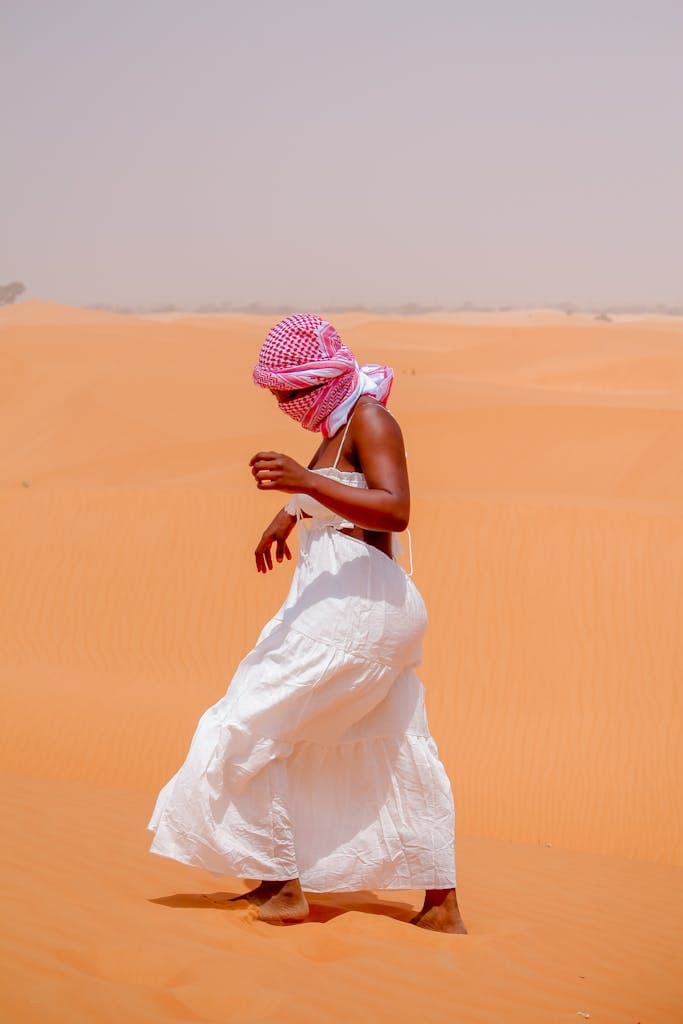 A woman in a white dress and pink headscarf walks in a desert landscape under clear skies.